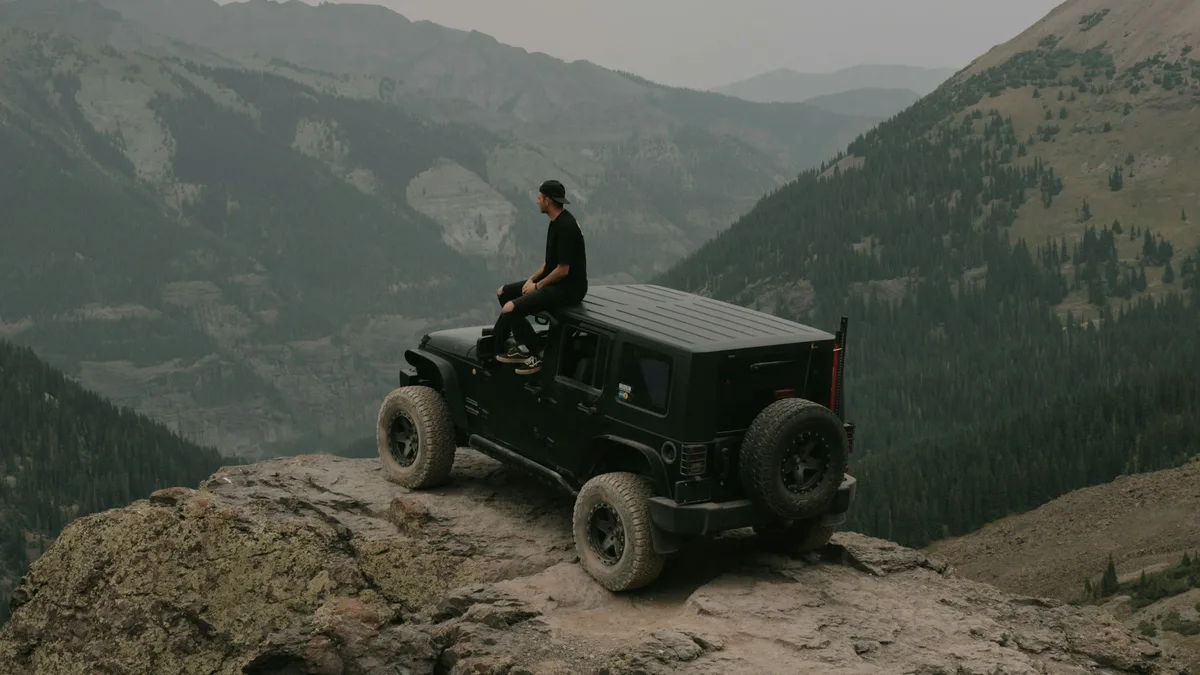 A black Jeep Wrangler 4x4 parked on a rugged rocky mountain overlook, with a rider taking in the backcountry view