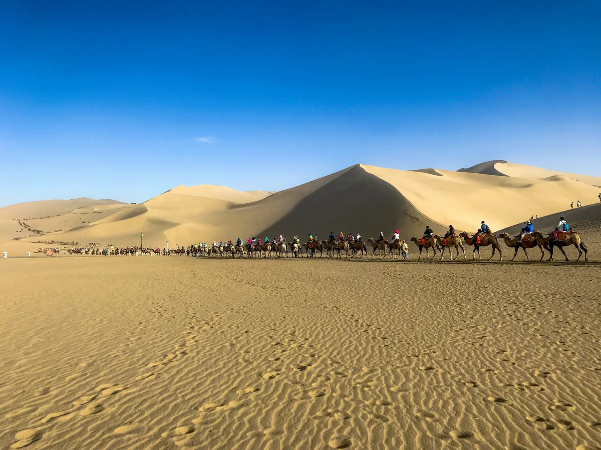 Camel riders crossing golden desert dunes near Dunhuang on China's ancient Silk Road