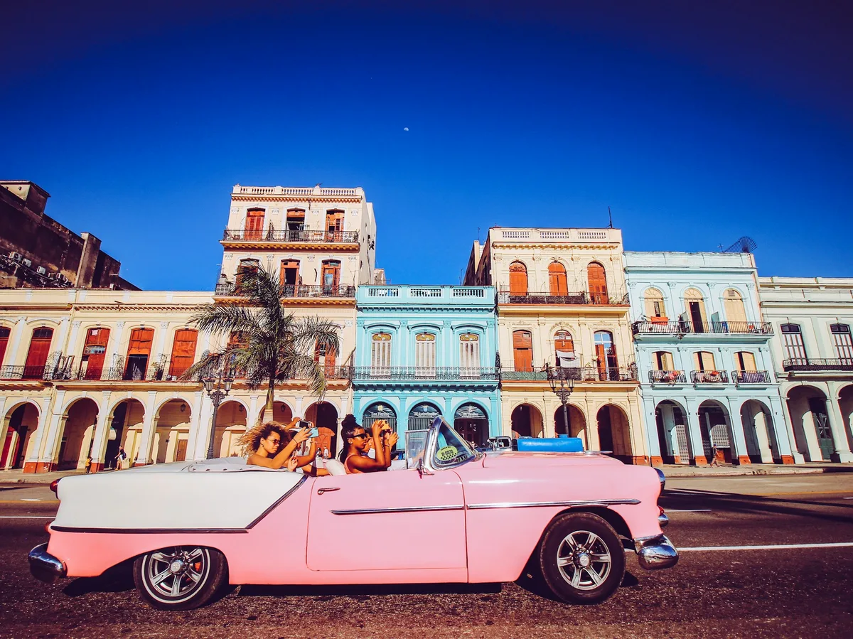 A pink vintage convertible with passengers driving past colourful colonial buildings in Havana
