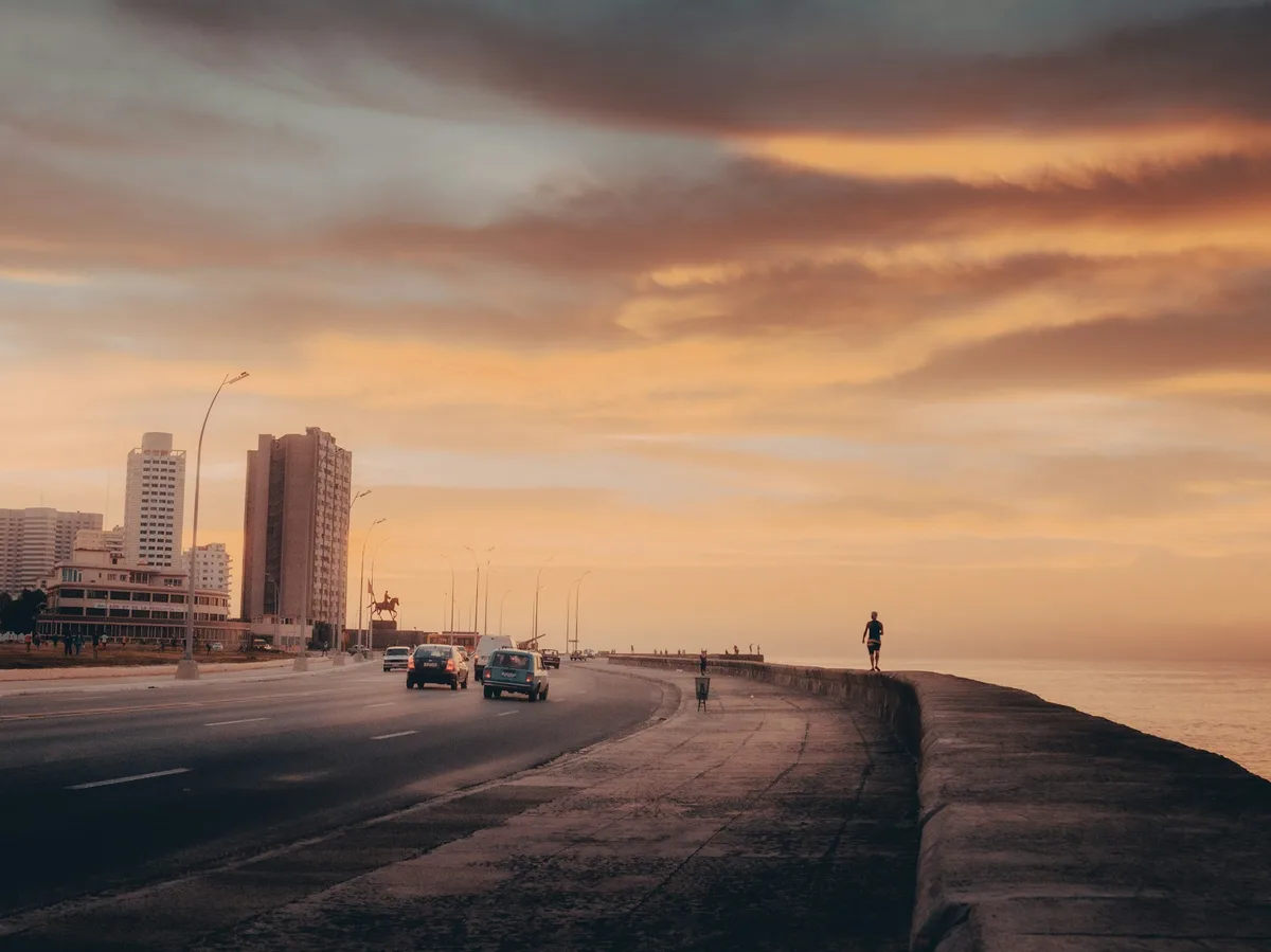 Sunset along Havana's Malecón seawall, with traffic on the road and a lone figure walking at the water's edge