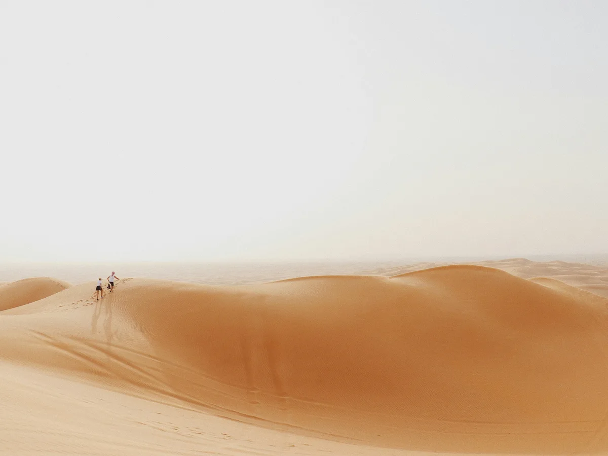 Two people walking up a large red sand dune in the Dubai desert