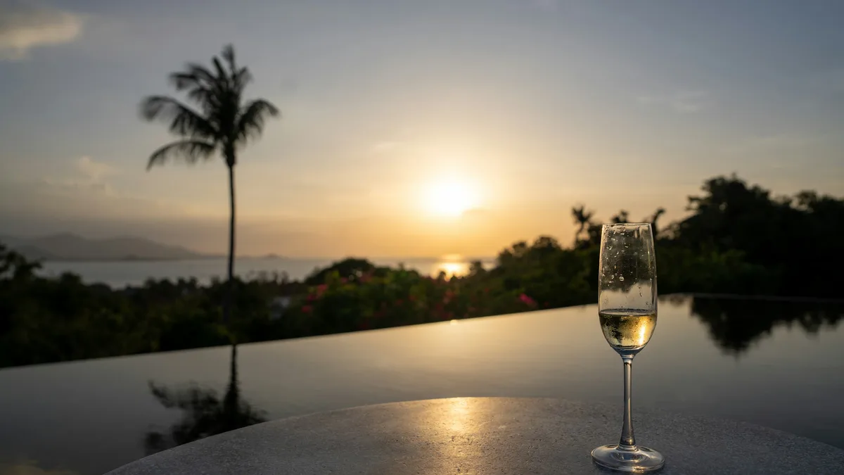 A glass of champagne on a table beside a tropical infinity pool at sunset, with palm trees and the ocean in the distance