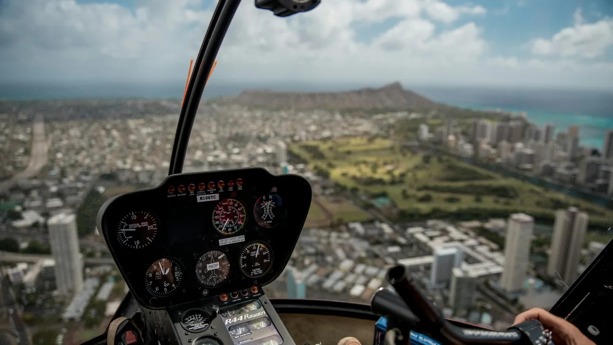 View from inside a helicopter cockpit over Honolulu, with Diamond Head crater and the turquoise coastline in the distance
