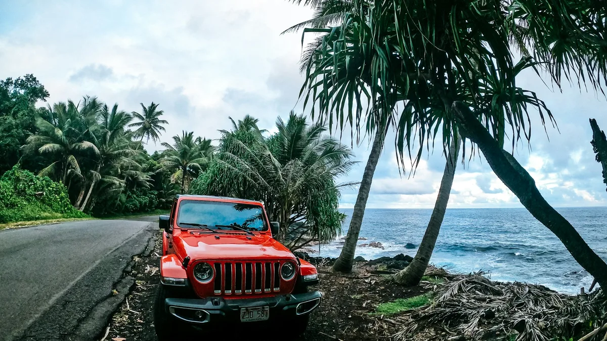 A red Jeep Wrangler parked on a palm-lined coastal road in Hawaii, with the ocean just beyond the trees