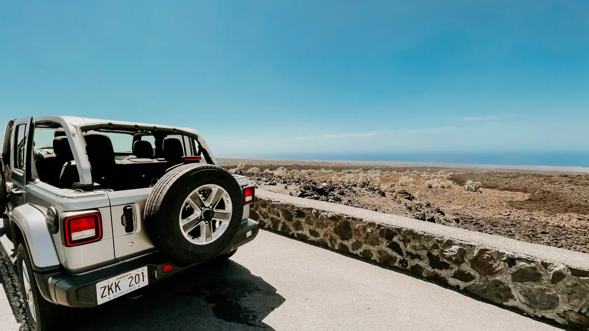 An open-top Jeep Wrangler parked along Hawaii's coast, with the ocean visible beyond a lava-rock wall