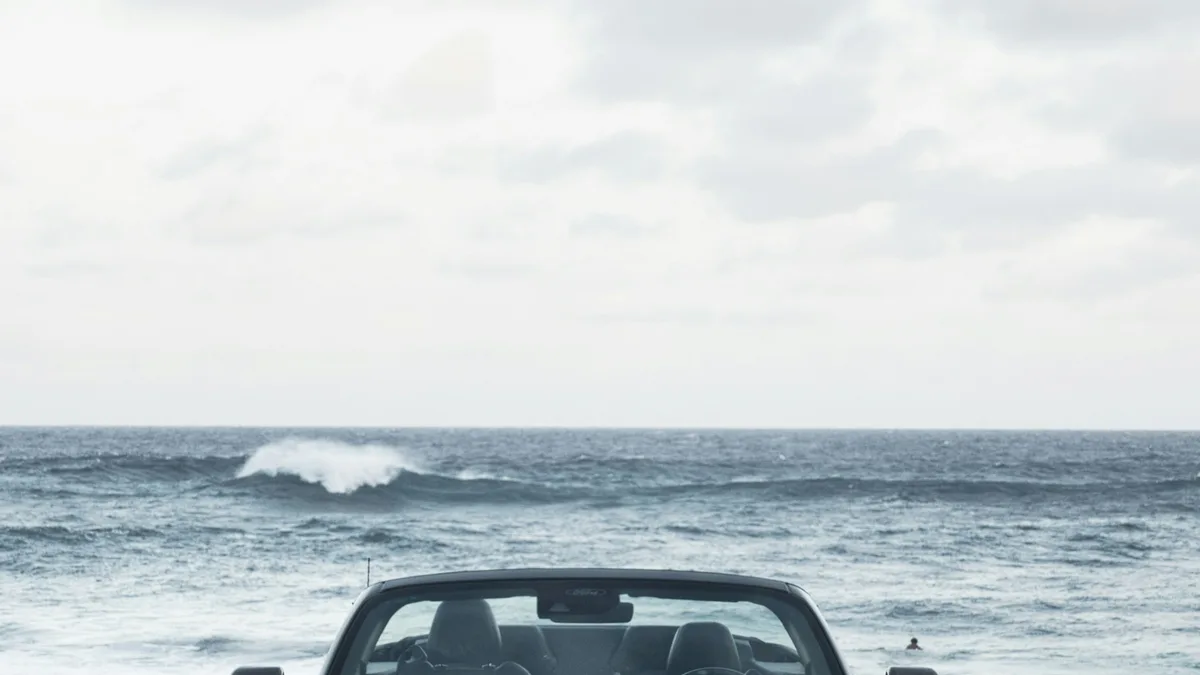 A convertible Ford Mustang with a Hawaii license plate parked facing the ocean, with waves rolling in behind it
