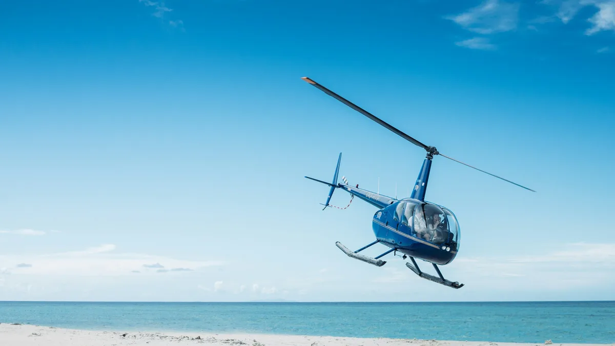 A small helicopter flying low over turquoise coastal water near a sandy beach