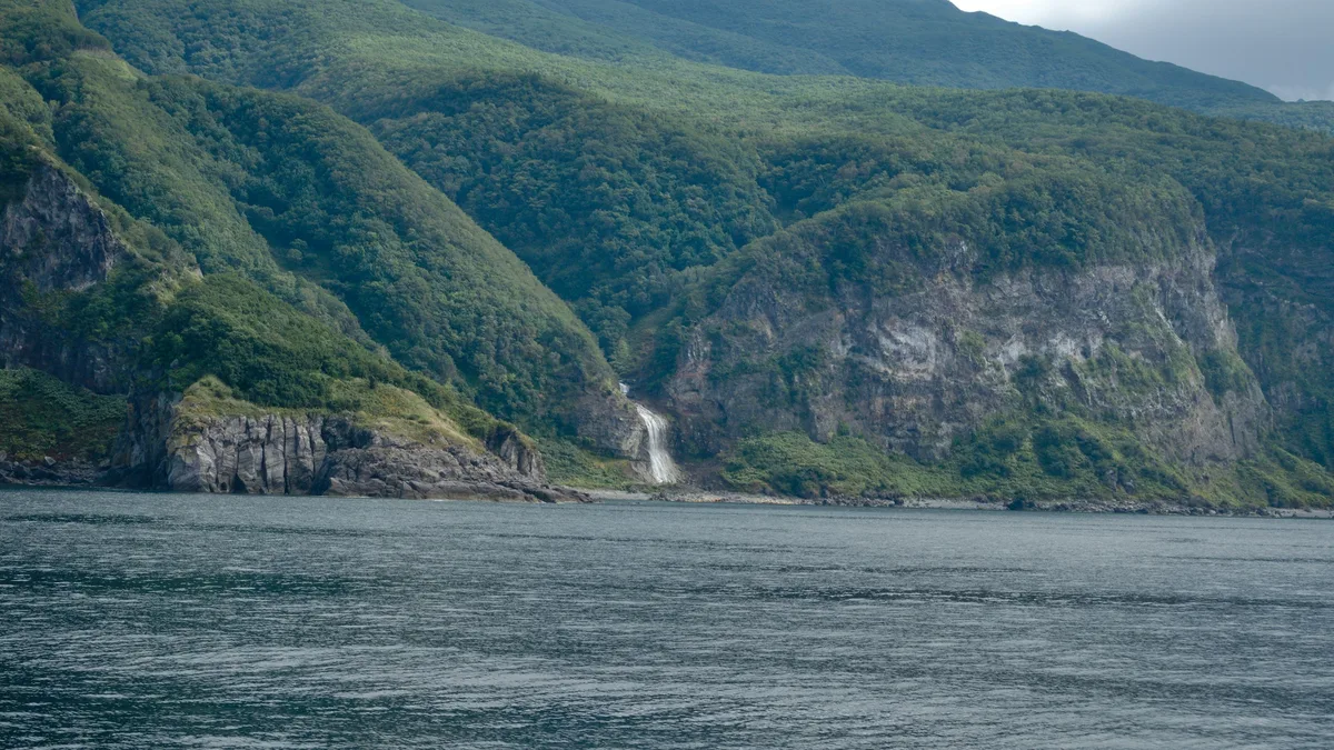 A waterfall cascading down a forested mountainside into a calm fjord